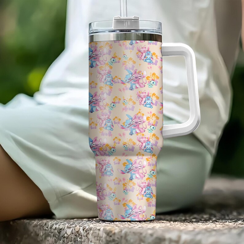 Puede incluir: Un vaso grande y aislado con asa y tapa blancas. El vaso presenta un patr&oacute;n repetido de personajes de dibujos animados en rosa y azul, con detalles florales sobre un fondo beige. Incluye una pajita transparente.