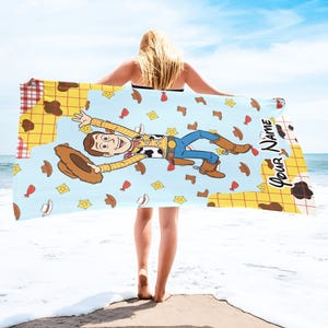 Puede incluir: Toalla de playa azul claro con una imagen de dibujos animados de Woody de Toy Story. La toalla tiene un fondo azul claro con Woody en el centro, rodeado de sombreros de vaquero, botas y insignias de sheriff. El texto "Your Name" est&aacute; impreso en la toalla.