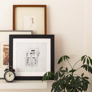 May include: A black framed print of a skeleton giving the middle finger and holding a cigarette. The print is on a white background and is hanging on a wall with other framed prints.