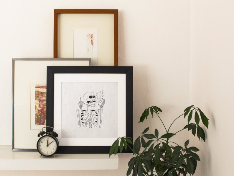 May include: A black framed print of a skeleton giving the middle finger and holding a cigarette. The print is on a white background and is hanging on a wall with other framed prints.