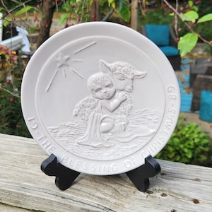 May include: A round white ceramic decorative plate featuring a raised relief design of an angel embracing a lamb under a star. The text "19-THE BLESSING OF CHRISTMAS-89" is embossed along the lower edge. The plate rests on a black plastic stand.
