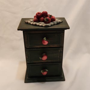 May include: A small wooden chest of drawers with a dark finish, featuring three drawers each equipped with a red apple-shaped knob. A square fabric cloth with a black and white checkered pattern sits on top, holding a cluster of small red apples.