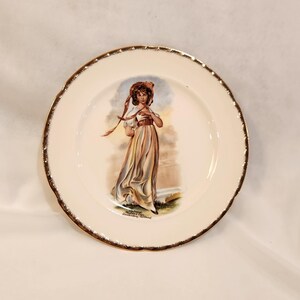 May include: A white ceramic plate with a gold rim features a colorful illustration of a woman in a long dress and a hat. The plate has the text "Souvenir of Connemara Ireland" printed on the bottom.