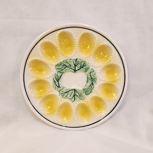 May include: A white ceramic egg plate with 12 yellow egg-shaped indentations. The center of the plate features a green leafy design.