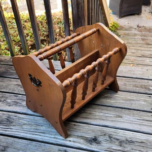 May include: A vintage wooden magazine rack with a warm brown finish. The rack features a curved design with decorative spindles and a handle. It is designed to hold magazines and newspapers.