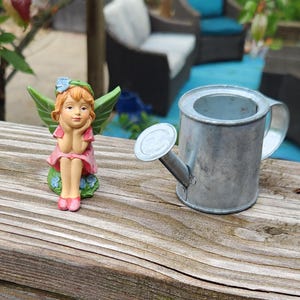 May include: A miniature fairy figurine with green wings, a pink dress, and a blue flower in her hair sits next to a small, galvanized metal watering can. The fairy is resting her chin on her hands. The watering can has a spout and handle.
