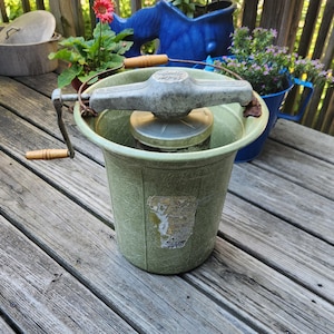 May include: A vintage light green ice cream maker with a textured fiberglass bucket, a metal hand crank mechanism with wooden handles, and a central metal canister. A weathered, partially torn paper label is attached to the front of the bucket.