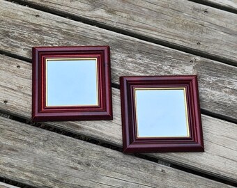 May include: Two dark red wood picture frames with gold trim and glass inserts. The frames are empty and are sitting on a wooden surface.
