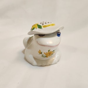 May include: White ceramic frog figurine with a yellow and green flower design. The frog is wearing a white hat with a yellow band and a yellow flower.