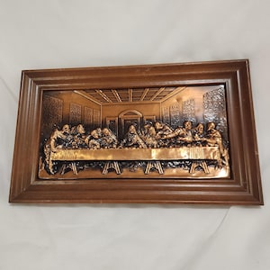May include: A rectangular copper-toned metal wall plaque depicting The Last Supper in relief, featuring a central figure seated at a long table with twelve individuals. The metallic scene is encased within a dark brown wooden frame with beveled edges.