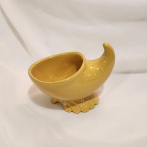 May include: A yellow ceramic cornucopia-shaped decorative bowl. The bowl has a curved, horn-like shape with a wide opening and a scalloped base. The surface is smooth and glossy, with a warm, golden-yellow color.