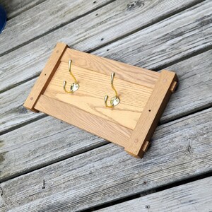 May include: A rectangular wall-mounted coat rack featuring a light brown oak wood frame with a natural grain pattern. Two polished gold-tone metal hooks are securely attached to the center panel, providing functional storage for hanging items.
