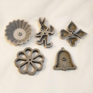 May include: Five gray metal cookie cutters in various shapes: a flower, a bunny, a bell, a four-leaf clover, and a scalloped circle. Each cutter has a hole for hanging or storage. These cutters are ideal for baking.