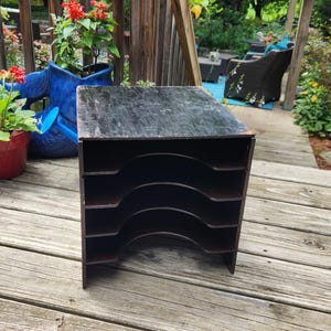 May include: A black wooden desktop document organizer featuring four horizontal shelves with curved cutouts on the front edges. The top surface shows visible wear and scuffing, while the interior shelves are finished in a dark brown tone.