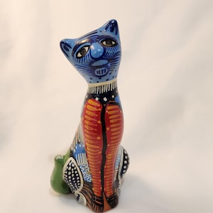 May include: A ceramic cat figurine with a blue head and body, featuring intricate patterns in red, orange, black, and white. The cat is sitting upright with its tail tucked in, showcasing a folk art style.