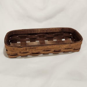 May include: A rectangular woven basket with rounded corners, constructed from thin, dark brown wooden splints. The basket features an open-top design with a visible grid-like weave pattern along the sides and a solid, flat base.