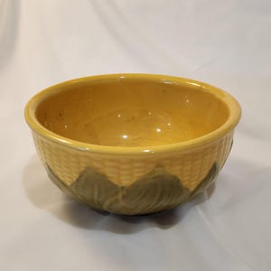 May include: A yellow ceramic bowl with a textured exterior resembling woven material. The bowl features a decorative pattern of green leaves around the base. The interior of the bowl is smooth and glossy.