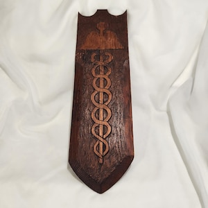 May include: A dark brown, wooden wall hanging with a carved medical symbol. The symbol is a staff with a serpent coiled around it. The top of the hanging has a carved design of a cross.