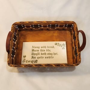 May include: A rectangular woven basket with brown leather handles. Inside the basket is a wooden base and a white tile with a poem printed in green. The poem reads: "Along with bread, Warm this tile, They'll both stay hot, For quite awhile."