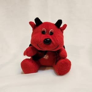 May include: Red plush toy devil with black horns and a black bow tie. The toy has a button that says "Press Here".