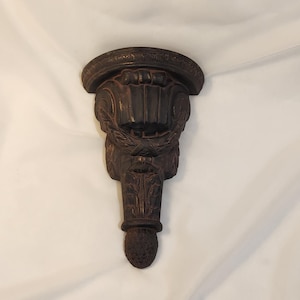 May include: A dark brown, ornate wall sconce with a decorative, carved design. The sconce features a rounded top, a central section with a laurel wreath, and a lower section ending in a textured, rounded shape. The overall design is reminiscent of classical architecture.