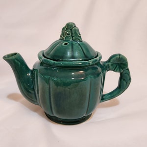 May include: A small, green ceramic teapot with a decorative lid and a leaf-shaped handle.