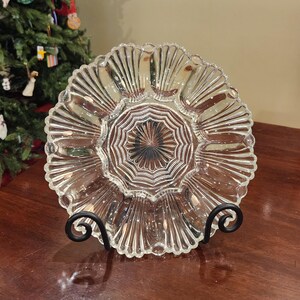 May include: A clear, decorative glass plate with a scalloped edge and a starburst design in the center. The plate is displayed on a black metal stand. The plate is likely used for serving or display.