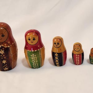 May include: A set of six wooden nesting dolls, each painted with a smiling face and decorative patterns. The largest doll is brown with star accents, while the others feature red, green, and black details. A classic folk art item.