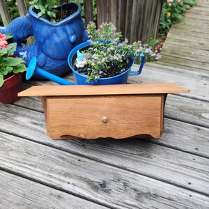 May include: A rectangular wooden wall shelf featuring a single drawer with a small, centered brass-colored knob. The shelf has a flat top surface and a curved bottom edge, showcasing a natural wood grain finish in a medium brown tone.