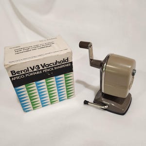 May include: A beige and dark brown Berol V-3 Vacuhold pencil sharpener with a hand crank and a metal base. Next to it is the original white cardboard box featuring blue and green triangular patterns and the text Berol V-3 Vacuhold.