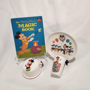 May include: A collection of Disney items including The Mickey Mouse Magic Book with a blue cover, a white plate featuring Mickey and friends with balloons, a white mug with Dumbo and Minnie Mouse, and a white bowl with a Minnie Mouse keychain.