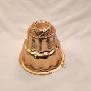 May include: A copper-colored Bundt cake mold with a decorative, fluted design. The mold has a ring for hanging and is made of a shiny, reflective metal. The cake mold is designed with a spiral top and a scalloped base.