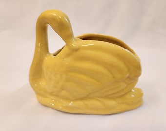 May include: A yellow ceramic swan planter. The swan has a long neck and a curved body. The planter has a small opening in the back for drainage.