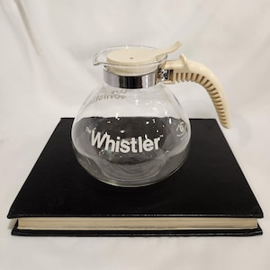 May include: A clear glass coffee carafe labeled The Whistler in white text. It features a cream-colored plastic lid and a matching ribbed handle attached with a chrome metal band. The carafe has a rounded body and a small spout for pouring.