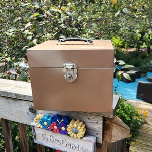 May include: A square, metallic bronze-colored storage box with a black handle on top and a silver-toned metal latch closure on the front. The box features a smooth, solid finish and is designed with a hinged lid for secure storage of various items.