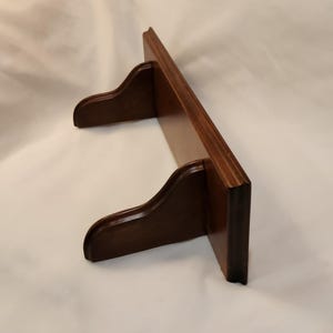 May include: A dark brown wooden shelf with two decorative brackets. The shelf is rectangular and appears to be made of solid wood. The brackets are curved and add a touch of elegance to the design. The shelf is designed for wall mounting.