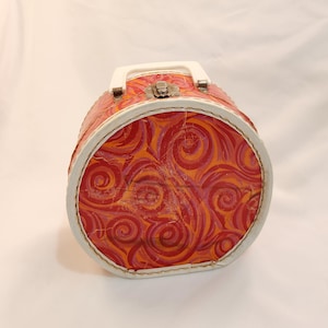 May include: A vintage, round, red and orange patterned case with a white handle and trim. The case has a metal clasp and is likely used for carrying small items. The design features swirling patterns in shades of red, orange, and pink.
