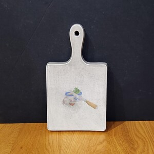 May include: A rectangular, light gray cutting board with a handle and a hole for hanging. The board features a small illustration of a knife, a blue ribbon, and a small green plant. The cutting board is made of a speckled material.