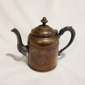 May include: A vintage copper teapot with a silver-plated handle and spout. The teapot has a rounded body and a decorative lid.