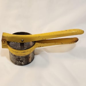 May include: A vintage potato ricer with a yellow handle and a perforated metal base. The handles are slightly curved and show signs of wear, suggesting age. The ricer is designed for mashing potatoes and other soft foods.