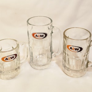 May include: Three clear glass mugs with handles. Each mug has a textured design and the A&W logo in orange, brown, and white. The mugs are of varying sizes, suitable for beverages.