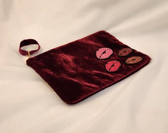 May include: A burgundy velvet clutch with embroidered lip designs in shades of pink and red. A matching burgundy choker with a gold clasp is also present. The clutch is rectangular and has a soft, textured appearance.