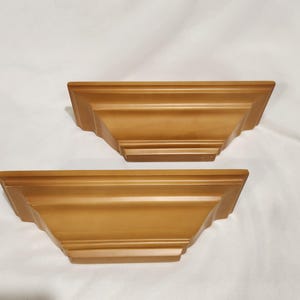 May include: Two identical wall-mounted shelves with a smooth, matte gold finish. Each shelf features a tiered, stepped design with a flat top surface and a tapered base, showcasing clean lines and a uniform, solid metallic-toned appearance.