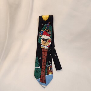 May include: Black novelty tie featuring a cartoon character dressed as Santa Claus in a chimney. The tie has a yellow moon at the top and a winter scene with a house and trees at the bottom. The tie is approximately 58 inches long.