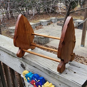 May include: A wooden towel rack with a natural finish. The rack has two upright, arrowhead-shaped supports connected by three horizontal dowels. The wood grain is visible, and the rack is set on a wooden deck.