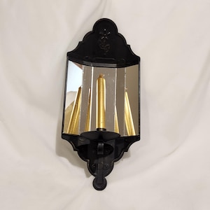 May include: A black metal wall sconce featuring a central mirror panel with a decorative scalloped top and bottom edge. A single gold-colored candle is positioned in the center of the sconce, held by a small circular black metal base.