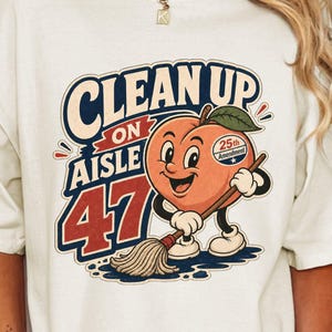May include: An ivory t-shirt featuring a graphic of a cartoon peach character holding a mop. The text reads "CLEAN UP ON AISLE 47" in blue and red, with a small sticker on the peach that says "25th Amendment". The design uses a retro color palette.