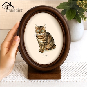 Puede incluir: Marco ovalado de madera marrón oscuro con un retrato de acuarela de un gato atigrado. El gato tiene rayas marrones y negras y ojos verdes. El marco se apoya en una base de madera. El texto "Family House Portraits" está en la parte superior.