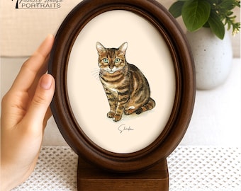 May include: An oval dark brown wooden frame with a stand containing a watercolor illustration of a brown tabby cat with green eyes. The name Shadow is written in black cursive script below the cat on a cream-colored background.