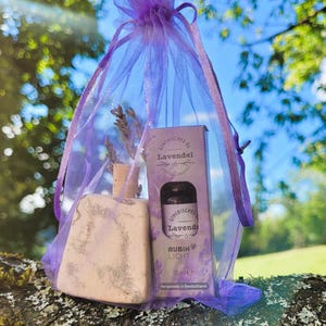 Puede incluir: Un juego de regalo con temática de lavanda en una bolsa de cordón morada transparente. El juego incluye una pequeña botella de cerámica con ramitas de lavanda seca y una caja de aceite esencial "Atherisches Lavendel".